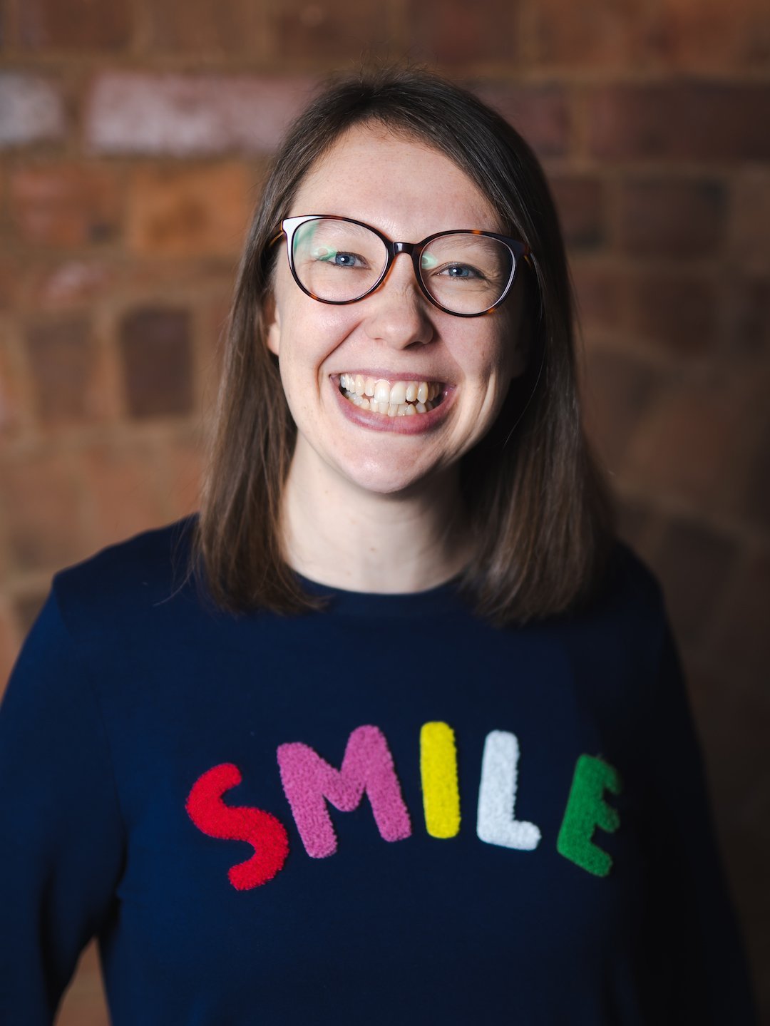 A white female with shoulder length brown hair and glasses is smiling. She is wearing a navy blue jumper with 'SMILE' written on the chest in different colours.