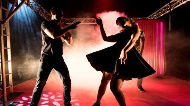 A girl and a boy stand silhouetted on stage, light up in red and blue. They are both dancing as if in a night club.