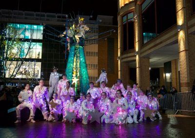 A group dressed in white with fairy lights and makeup pose around a metal frame decorated like a Christmas tree.