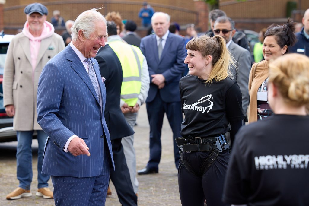 Prince Charles, an elderly white haired man wearing a blue suit, is laughing a talking to a woman wearing a black t shirt with a logo on reading 'CastAway.' 