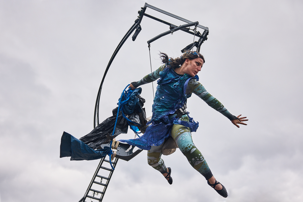 A woman in a dark blue tattered dress flies in the air, suspended from an aerial rig. She looks troubled and is holding a string of rubbish and paste waste.