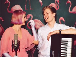 Fi and Alex stand in front of a green wall, patterned with flamingo wallpaper. Fi is a white woman with pink short hair. Alex is a white man with short blond curly hair. They are holding a keyboard and a micrphphone.