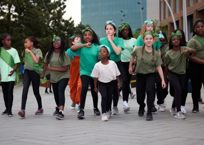 Young girls, all children, are wearing green t-shirts and black trousers, and are all wearing green headbands. Most of the children are looking ahead. Behind them is a woman in a blue t-shirt and orange overalls, who is shouting something.