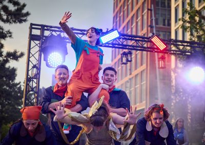 On the street, a six people have made a pyramid shape. On the bottom is two women in blue jumpsuits and orange headbands, and a women in a sparkly gold jumpsuit, with gold and silver sleeve. Two men, in blue jumpsuits and orange neckties are behind, and carrying a woman wondrously looking and pointing to the sky, wearing a blue t-shirt and orange overalls.