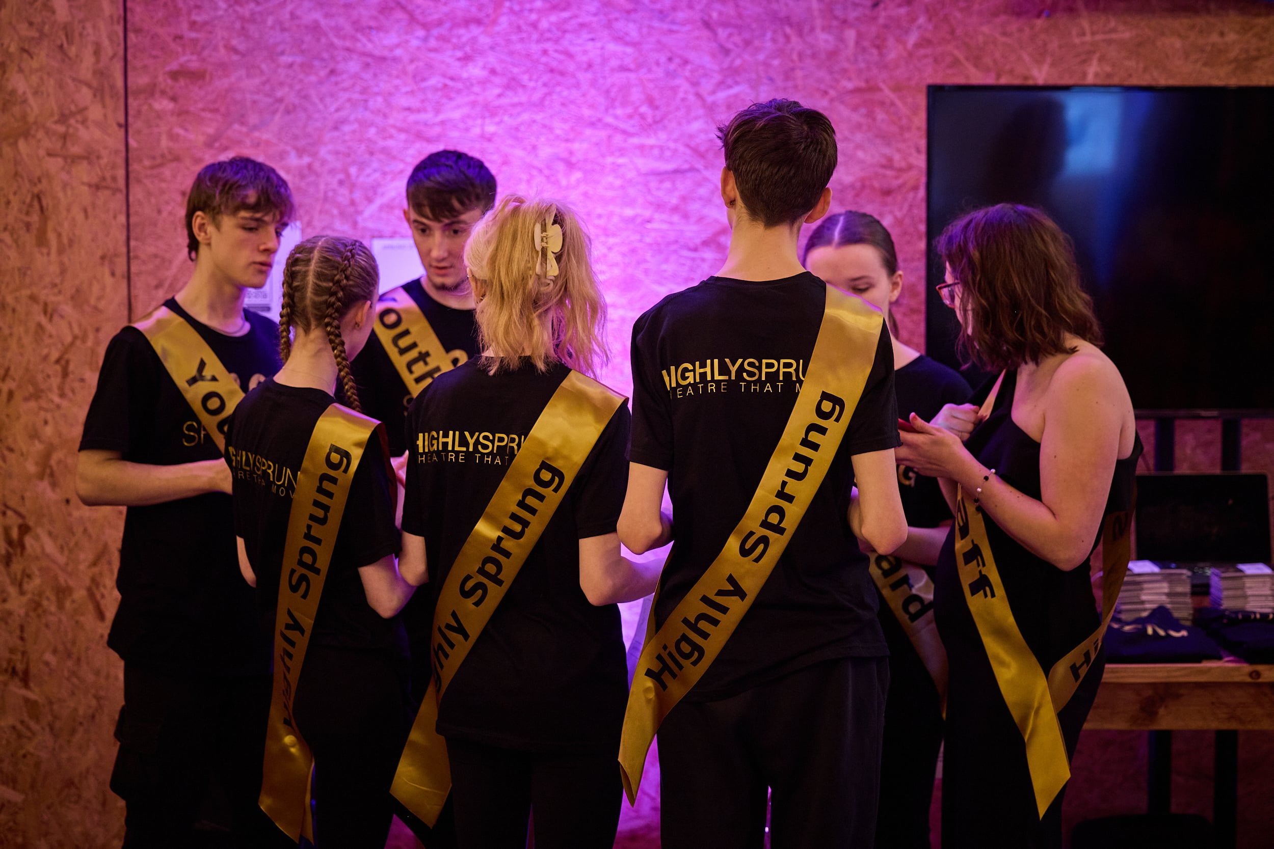 A group of teenage performers stand on stage. They are wearing suits and all holding large pieces of paper in different directions,as if reading.