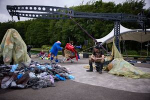 An outdoor performance next to the canal. There is an aerial rig, with a man sitting next to it and a big pile of rubbish. Behind the rig are 2 performers dressed in bright red and blue coats, messing around in a shopping trolley.