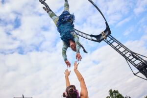 A woman hangs upside down from a metal rig, reaching her hands out to a woman who stands on the ground beneath her.