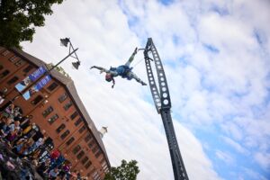 A woman in a dark blue dress is suspended upside down from an aerial rig in the air, against a blue sky.