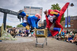 2 performers in bright red and blue coats run along next to a shopping trolley, kicking up and jumping in the air.