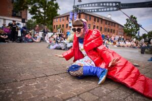 An outdoor performance -A woman in a shiny red coat and sunglasses crouches down on the floor laughing.