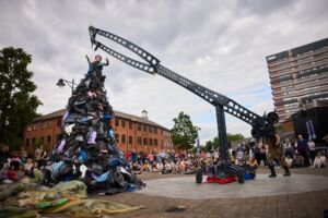 An outdoor performance. In the middle of a square, there is a tall aerial rig and a woman wearing a long dress made of rubbish and plastic bags.
