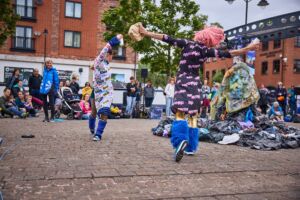 2 women in colourful, patterned dresses dance, moving their arms up through the air. They are holding pieces of rubbish.