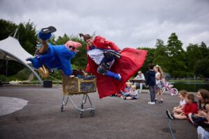 2 performers in bright red and blue coats run along next to a shopping trolley, kicking up and jumping in the air.