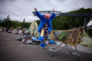A woman in a pink wig, gold leggings and blue furry coat jumps in the air next to a shopping trolley.