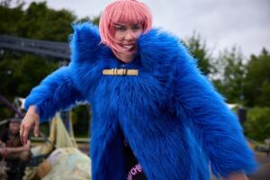 A female with pink bob and a fringe is smiling at the camera. She wears a blue furry coat.
