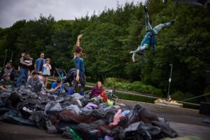 An outdoor performance, in the foreground with a big pile of rubbish. Behind it, a woman stands and reaches up in the air towards another performer who is suspended from an aerial rig.
