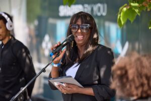 Sandra Godley stands smiling, talking into a microphone. She is outdoors on a sunny day, under some trees. Sandra is a black middle aged woman with shoulder length brown & black hair. She wears a black blazer and is in sunglasses.