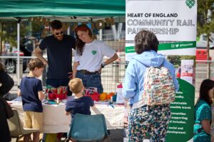 An outdoor stall with a table full of craft and creative activities & resources. There is a sign reading 'Heart of England Community Rail Partnership.' 2 small boys stand next to the stall.