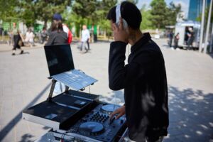 A DJ wears headphones and stands next to a deck and laptop. He is outdoors in a square. The DJ is black, with short hair.