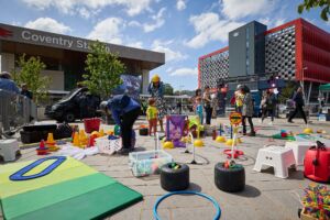 Playhem at the Promenade, Coventry Transport Museum, credit Andy Moore An outdoor square. In the background is Coventry Station, against a cloudy blue sky. In the foreground is a collection of plastic toys and signs, all related to travel and transport. There are some adults and children stood nearby.