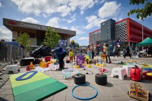 An outdoor square. In the background is Coventry Station, against a cloudy blue sky. In the foreground is a collection of plastic toys and signs, all related to travel and transport. There are some adults and children stood nearby.