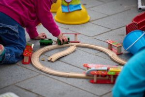 A child plays with a wood train and train track. It is outdoors on top of some concrete paving slabs.