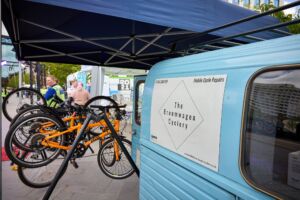 A close up of a sign on a blue van, reading 'The Broomwagon Cyclery.' In the background you can see a collection of bikes.