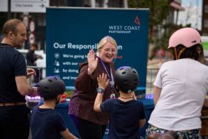 A woman laughs and high fives a child who wears a cycling helmet, both standing in front of an 'Avanti West Coast' sign. The woman is white, in her thirties, wears glasses and has blonde hair that is tied back.