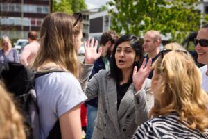 Zarah Sultanah, a South Asian MP in her 20s, stands talking to a group. She wears a grey suit, and has shoulder length black hair. She is holding her hands up in the air as she speaks.