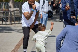 A crowd watches John Bernard, a rapper, as he performs in an outdoor square outside Coventry station. In front of him a toddler is dancing.
