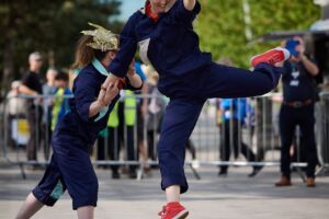 In the street, two people are dancing together, both wearing dark blue jumpsuits and red converse shoes. One woman, with an orange headband, has jumped in the air, with her left leg suspended at a right angle position. The other woman, who is wearing a gold headpiece, is standing behind, holding her hand