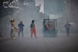 There is a street covered in smoke, and along it is a vehicle made out of metal pipes, and three golden wheels at the front. On the street, there are three women and one man. The woman on the left is wearing a sparkly gold outfit and carrying a red wheel prop. Next to her is a woman in a blue jumpsuit. Next to her is another woman wearing a light-blue t-shirt and a bright orange jumpsuit, who is also carrying the red wheel prop. The man is standing on the right, and is wearing a denim jumpsuit.