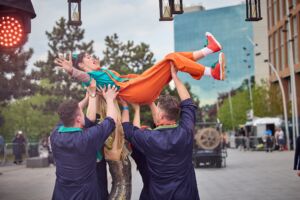 In the street, four people dressed in blue jumpsuits, and one woman in a sparkly gold jumpsuit are lifting a woman in a blue t-shirt and orange overalls. She is looking excited and surprised, and a red colour LED and candles are suspended above.