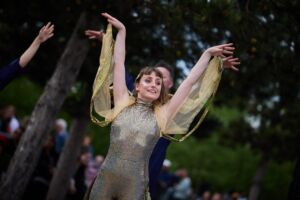 A woman in a gold, sparkly jumpsuit has her arms lifted upwards. There are fabrics attached to the underneath the armpit section of the jumpsuit and the fingers, creating a wing like image. Behind her, men in blue jumpsuits are replicating her movements.