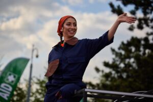 Outside, a woman in a dark blue jumpsuit with brown pockets, and wearing a red bandanna is signalling to the right. She is looking to the right with a smile.