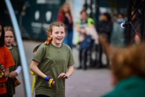 A young girl with brown hair is wearing a dark green t-shirt and has a multicoloured ribbon in her left hand. Standing behind her are a few more young children, who are all inquisitively looking at someone standing in front of them.