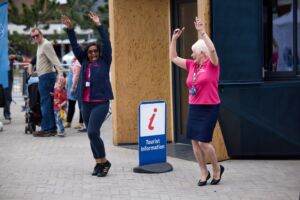 2 woman are dancing outdoors next to a sign that reads 'Tourist Information.' Both have one legs slightly bent and lift both arms into the air, pointing upwards and laughing. They are both middle aged & wear bright blue and pink clothing. One is white, with a short white pixie cut. The other woman is South Asian, has short black hair and wears sunglasses.