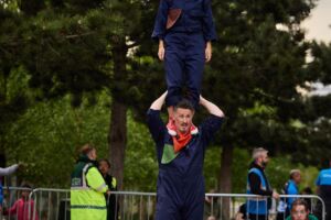 In the streets, a man in a dark blue and green jumpsuit and red trainers is holding up a woman on his shoulders. She is wearing a dark blue and green jumpsuit. Behind her, a man in a green jumpsuit is crouching down. Men in blue high visibility jackets are standing behind a barrier.