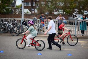 2 children ride bikes around a car park, in an area marked by cones. They are both white, in colourful clothing and are wearing helmets. There is a white middle aged woman with short hair who is standing nearby and providing instruction.
