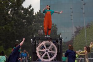 On the streets, a woman in a blue t-shirt and orange overalls, is standing on an old-fashioned looking vehicle, made from metal pipes and silver wheels. On the ground, and looking at the woman on the vehicle are three people in dark blue jumpsuits and red converse shoes, and a woman in a gold, sparkly jumpsuit.