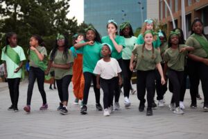 Young girls, all children, are wearing green t-shirts and black trousers, and are all wearing green headbands. Most of the children are looking ahead. Behind them is a woman in a blue t-shirt and orange overalls, who is shouting something.