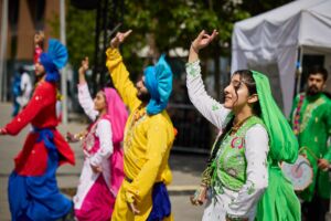 A group of bhangra dancers perform outside. In the foreground is a south asian woman, with a decorative green, pink and white clothing, lifting one arm into the air.