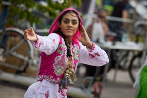 A south asian bhangra dancer performs outside. She is wearing decorative pink, white and gold clothing. One hand is behind her head as she reaches the other out to the side.
