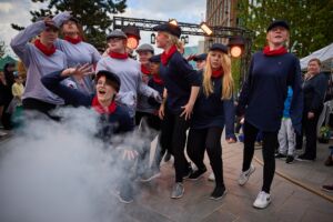 Young girls are in the streets, dressed in either dark blue or light grey tops, wearing matching hats, and all are wearing red neckties. Smoke is coming out from a machine, and all the girls are looking past it into the distance. An audience is watching from behind.