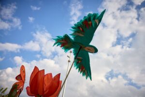 A turquoise bird kite with red, green and white feathers is flying in the sky. Next to it, is a red tulip kite, and a red and green butterfly.