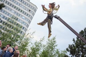 A woman in a gold jumpsuit sparkly jumpsuit is suspended in the air on an aerial flying machine. Watching below are an audience looking up, very impressed by what they are witnessing.