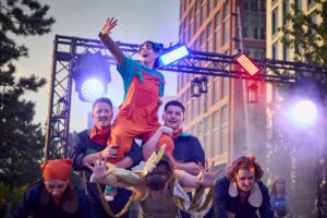 On the street, a six people have made a pyramid shape. On the bottom is two women in blue jumpsuits and orange headbands, and a women in a sparkly gold jumpsuit, with gold and silver sleeve. Two men, in blue jumpsuits and orange neckties are behind, and carrying a woman wondrously looking and pointing to the sky, wearing a blue t-shirt and orange overalls.