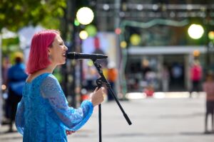 A jazz singer performs outdoors on a sunny day. She is a white woman with short pink hair, wearing a blue dress.