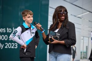 Sandra Godley stands interviewing a teenager, and they are both smiling out cheekily to the crowd. Sandra is a black middle-aged woman, with long black & brown hair, wearing sunglasses. The teenager is white, with short blonde hair, and wears a blue, black and white jacket.
