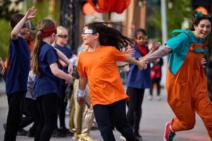 A female child wearing a bright orange t-shirt, black trousers and silver, opaque glasses is excitedly dragging by the hand a woman in a blue t-shirt, orange overalls and curly hair. Behind her, other children are wearing dark blue t-shirts and silver opaque glasses, and are dancing alongside a woman in a sparkly golden jumpsuit.
